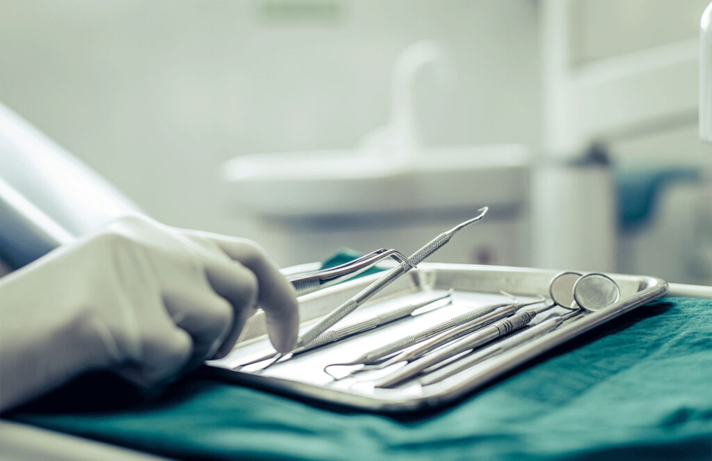 Close-up view of a gloved hand selecting dental instruments from a tray in a dental clinic, focusing on the precision tools used for dental treatments, against a soft green and white background.