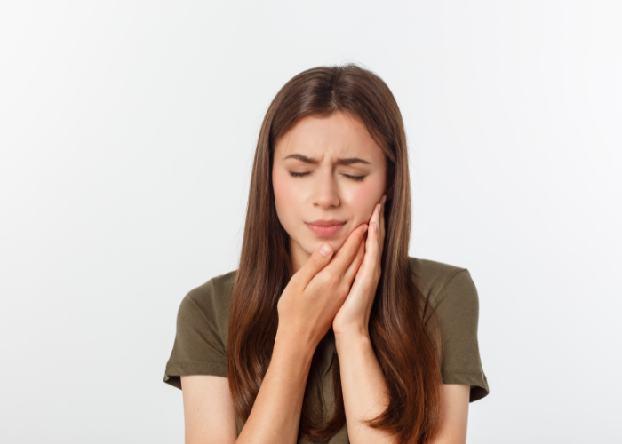 The image shows a young woman with long brown hair holding her cheek in pain, indicating discomfort or a toothache, against a plain white background.