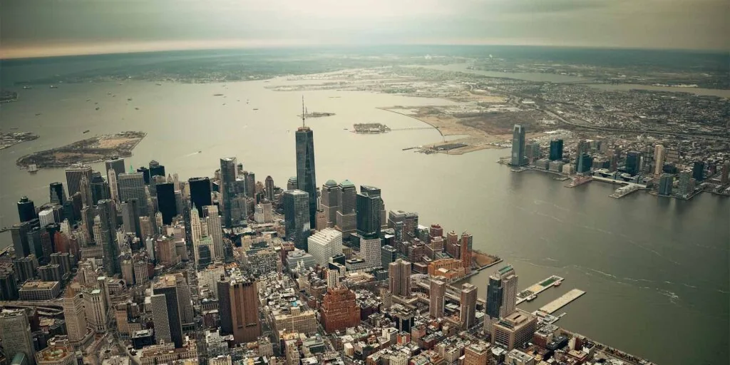 The image shows an aerial view of Lower Manhattan in New York City, with the One World Trade Center prominently in the center, surrounded by skyscrapers, and the Hudson River and New Jersey visible in the background.