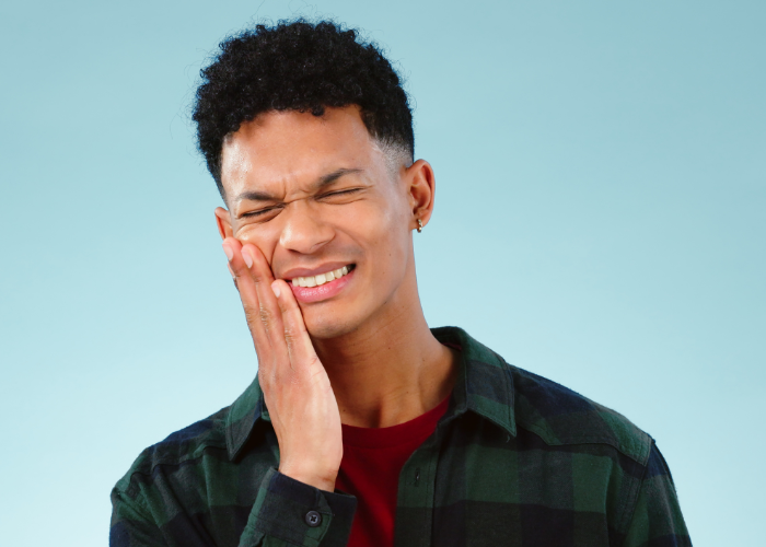 The image shows a young man grimacing and holding his cheek in pain, indicating a toothache, against a light blue background.