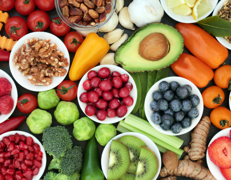 The image shows a colorful assortment of fresh fruits, vegetables, nuts, and other healthy foods, including avocado, blueberries, kiwi, tomatoes, bell peppers, garlic, walnuts, cranberries, and broccoli, arranged on a dark background.